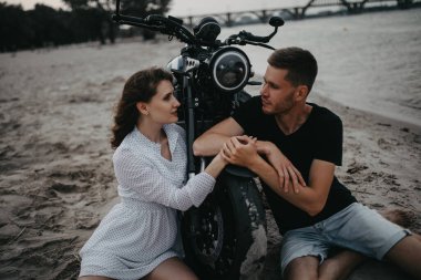 Birbirine aşık çiftler, motorsikletin yanındaki kumsalda oturup el ele tutuşurlar. Yakın çekim..