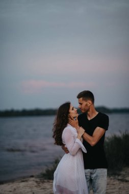 Birbirine aşık bir çift kumlu sahilde su ve gökyüzü geçmişine karşı durup sarılıyor..