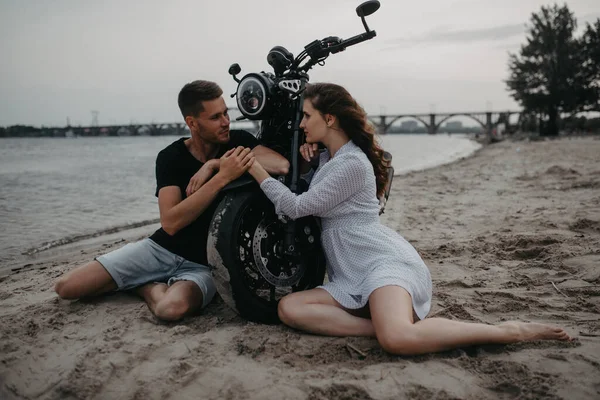 Birbirine aşık çiftler oturur ve kumsalda motosikletin yanında el ele tutuşurlar..