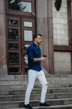 Genç adam elinde bir fincan kahveyle merdivenlere çıkıyor ve modern binanın arka planına dayanıyor..