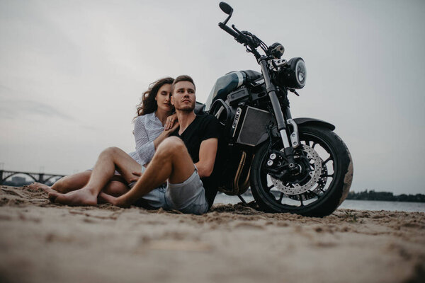 Couple in love sits and hugs on sandy beach near motorcycle. Bottom view.