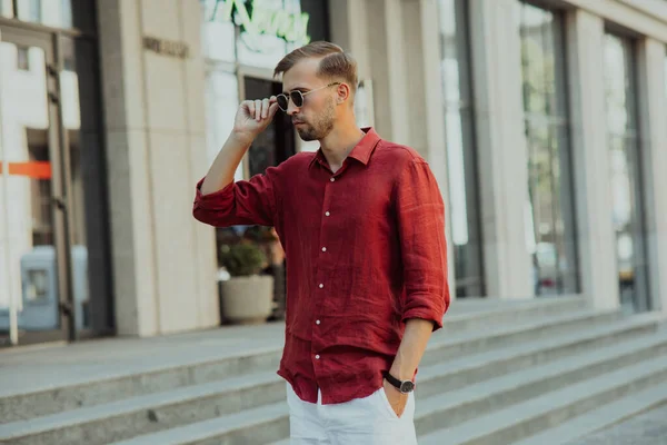 Young man in pants, shirt and sunglasses stands on street against background of building.