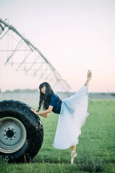 Japanese ballerina in white skirt stands and performs swallow pose in field on farm near the wheel of agricultural sprayer.