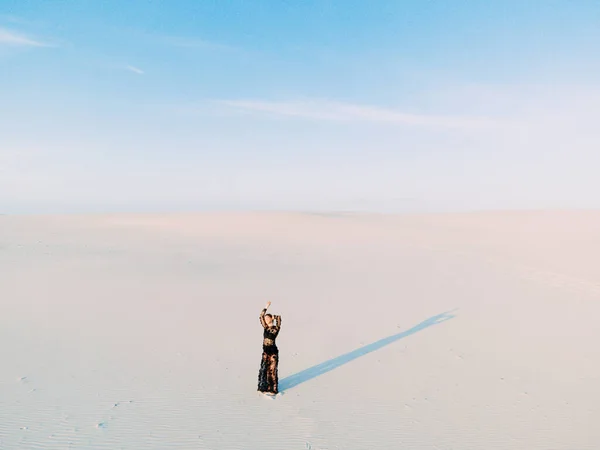 Woman in lace black dress stands in desert and casts a shadow on white sand. View from drone.