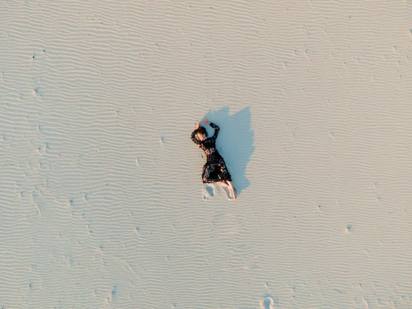 Woman in lace black dress lies in desert and casts a shadow on white sand. View from drone directly above.