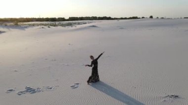 Dantelli siyah elbiseli kadın çölde dans eder ve beyaz kumda gölge bırakır. Yukarıdaki ve etrafındaki İHA 'dan görüntüle.