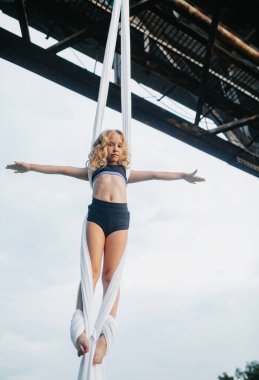 Child girl aerialist performs acrobatic tricks on hanging aerial silk attached to the bridge support against background of sky. clipart
