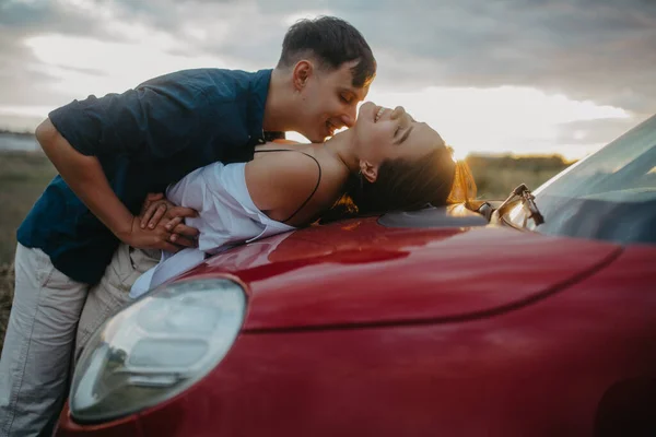 若いですカップルで愛のキスと包含するオン赤車Bonnetで草原で日没 — ストック写真