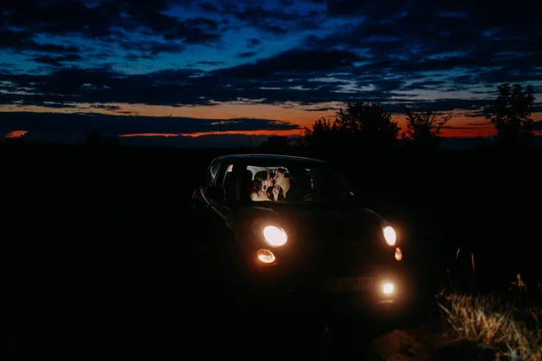 Young couple in love sits inside of car with glowing headlights and kisses against night sky background on meadow.