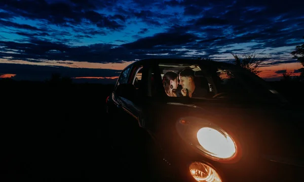 Young couple in love sits inside of car with glowing headlights and embraces against night sky background on meadow.