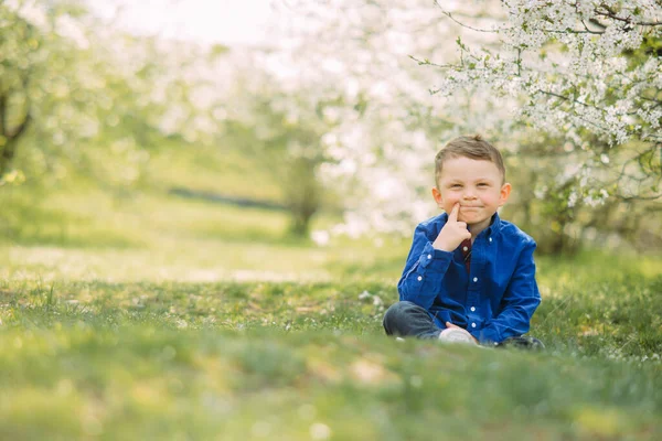在开着花的春天花园的背景下 快乐的小男孩坐在阳光下的草地上 — 图库照片