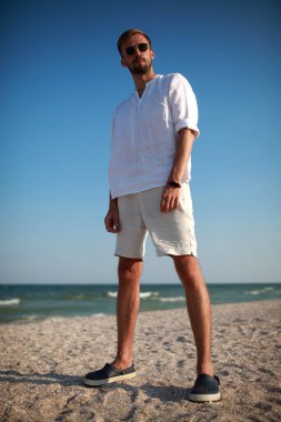 Güneş gözlüklü genç adam deniz ve gökyüzü arka planına karşı sahilde duruyor. Alt görünüm.