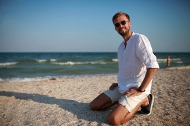 Güneş gözlüklü genç adam sahilde oturur ve gülümser. Deniz ve gökyüzüne karşı..