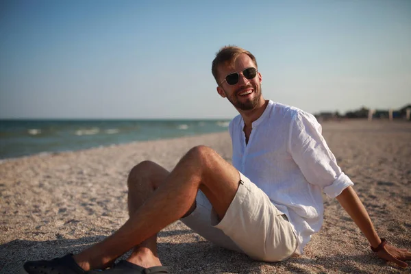 Joven Gafas Sol Sienta Sonríe Playa Contra Mar Cielo Fondo — Foto de Stock