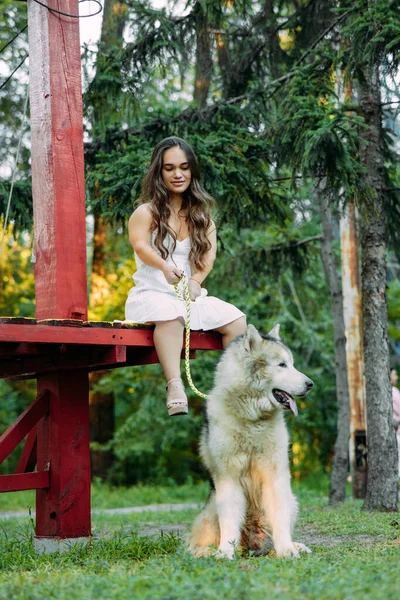 患有先天疾病的年轻女子侏儒坐在马拉穆特犬旁边的长椅上 一边在公园里散步一边系着皮带 — 图库照片