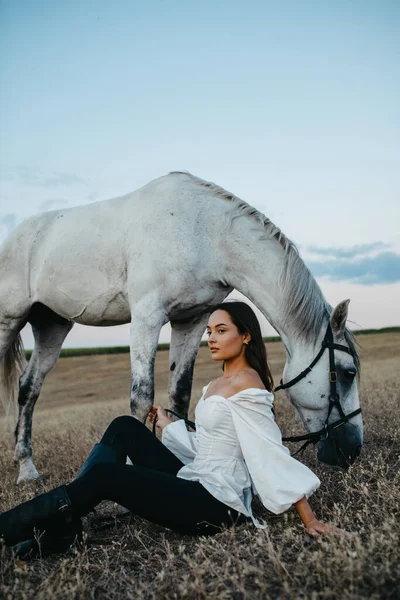 若い女性は牧草地で灰色の馬の近くに座っています — ストック写真