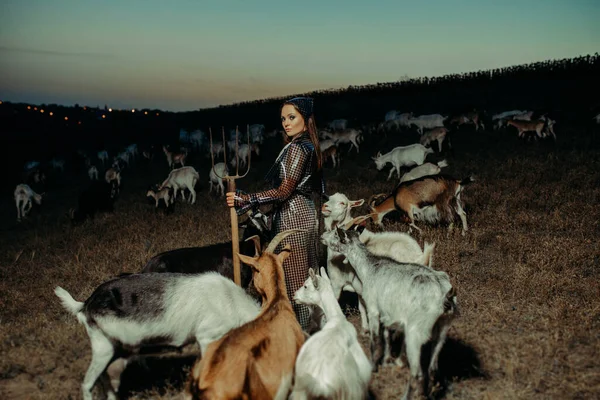 Handsome woman farmer stands in raincoat with pitchfork in her hand among a herd of goats on pasture in the night.