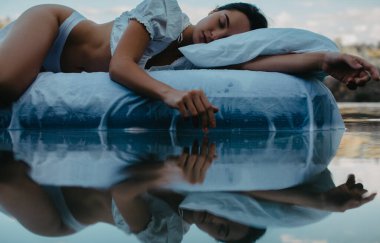 Young woman sleeps on a mattress with white bedding floating in the water with reflection. clipart