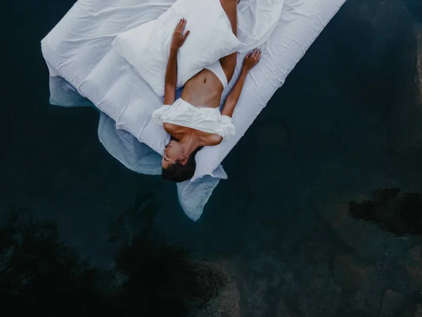 Young woman sleeps on a mattress floating in the water with white bedding. Top view.