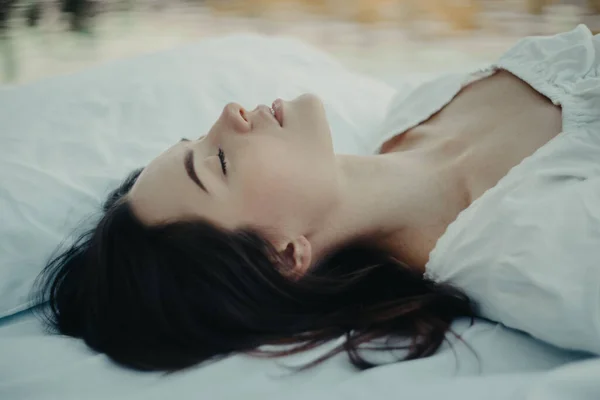 Young woman sleeps on a mattress with white bedding against background of water. Closeup.