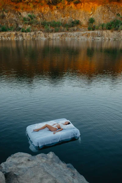Young woman sleeps on a mattress floating in the water with white bedding near rocks.