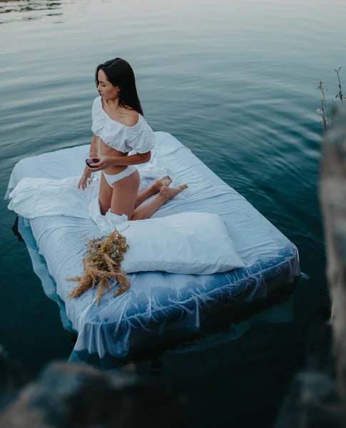 年轻女人躺在漂浮在水里的床垫上放松下来 手上拿着白纱和酒杯 — 图库照片