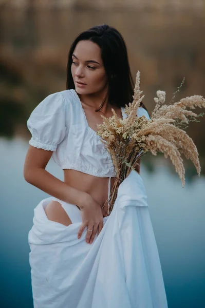 年轻的女人站在白色的床单上 背靠着湖水 手里拿着花束 — 图库照片