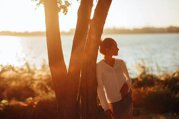 若い女性は日没時に川の背景を背景に木の近くに立つ — ストック写真
