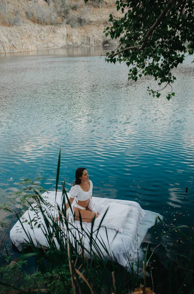 Young woman rests on a mattress floating in the water with white linens among plants.
