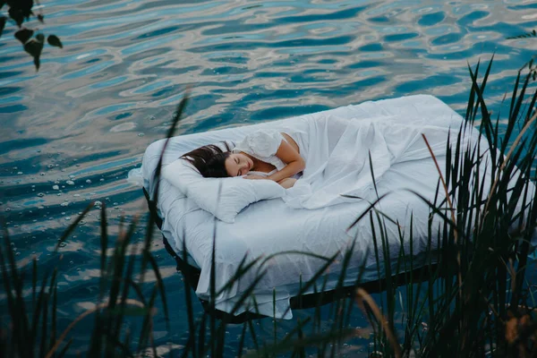 若い女性は植物の間で白いリネンと水の中に浮かぶマットレスの上で眠る — ストック写真