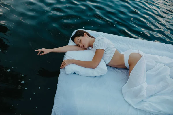 Young woman sleeps on a mattress floating in the water with white bedding. Top view.