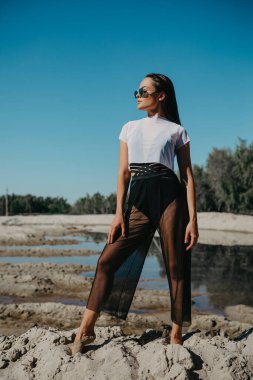 Güneş gözlüklü güzel kadın kumsalda mavi gökyüzüne karşı su kenarında poz veriyor..