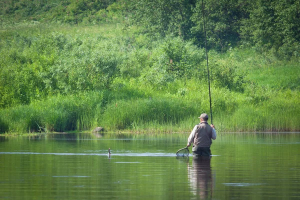 夏天在湖上钓鱼 一个人把一条鱼从河里拉了出来 手里拿着鱼竿和渔网 美丽的夏日风景 很多绿色的植被 反映在水中 — 图库照片