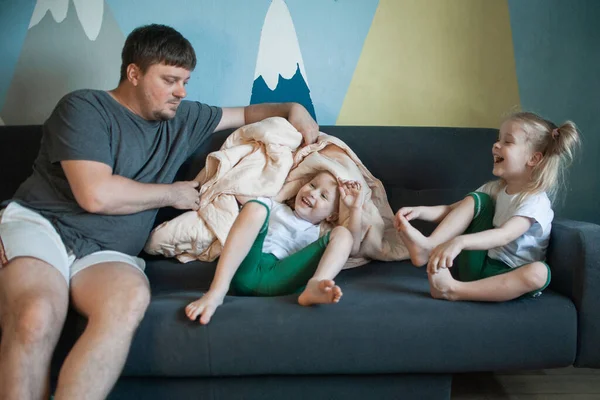 Dad plays hide and seek with his daughters under the covers. Dad and daughters play in a house with a blanket
