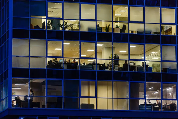 Windows of glass office building, lighting and working people within
