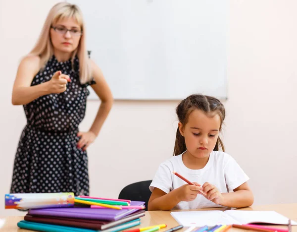 a strict teacher and a little student. Little girl is sad at the desk in the classroom