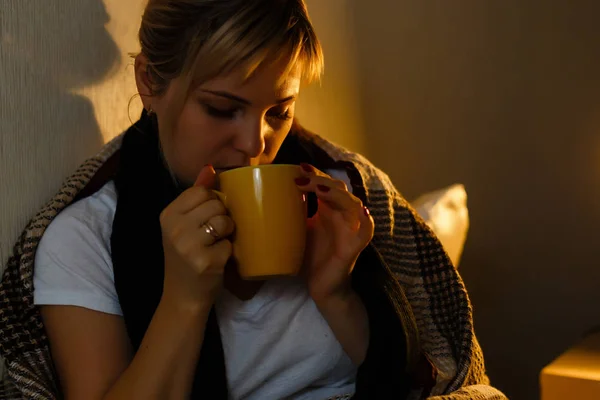 Sick woman with cup of tea. Closeup image of young frustrated sick woman in knitted scarf holding a cup of tea while lying in bed. feeling bad.