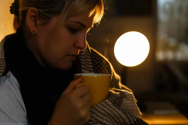 Sick woman with cup of tea. Closeup image of young frustrated sick woman in knitted scarf holding a cup of tea while lying in bed. feeling bad.