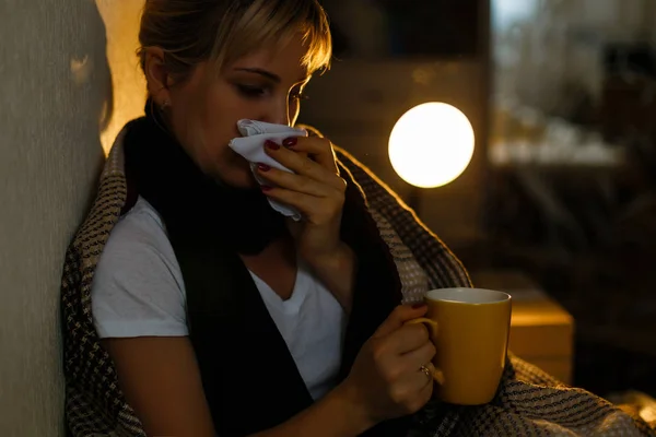 Sick woman with cup of tea. Closeup image of young frustrated sick woman in knitted scarf holding a cup of tea while lying in bed. feeling bad.