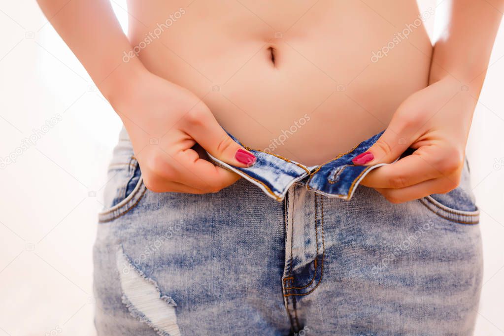 Weight gain woman getting dressed wearing jeans. Diet concept - closeup of women hands unable to close the pants due to gaining fat on hips. Fashion girl putting pants on.