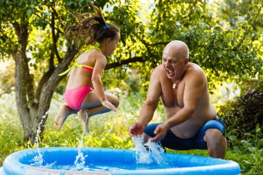Grandfather with granddaughter playing in an inflatable pool in the garden near the house clipart