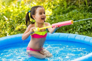 Happy little girl playing with water pump in outdoor inflatable pool on hot summer day. Child water toys. Water pump for playing with water clipart