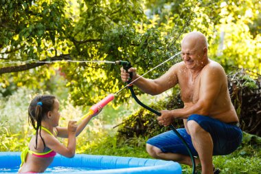 Grandfather and granddaughter pour each other with water in an inflatable pool in the garden near the house clipart