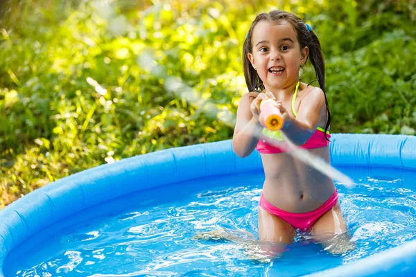 Happy little girl playing with water pump in outdoor inflatable pool on hot summer day. Child water toys. Water pump for playing with water
