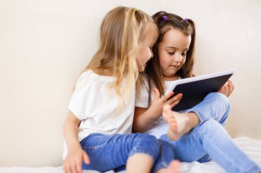 İki küçük kız eğleniyor dijital tablet kullanarak yatağın üstüne oturuyor