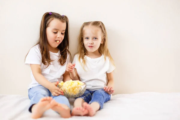 Movie night. Two young beautiful girls sitting on a white couch discussing a movie while holding remote control and eating popcorn at pajama party
