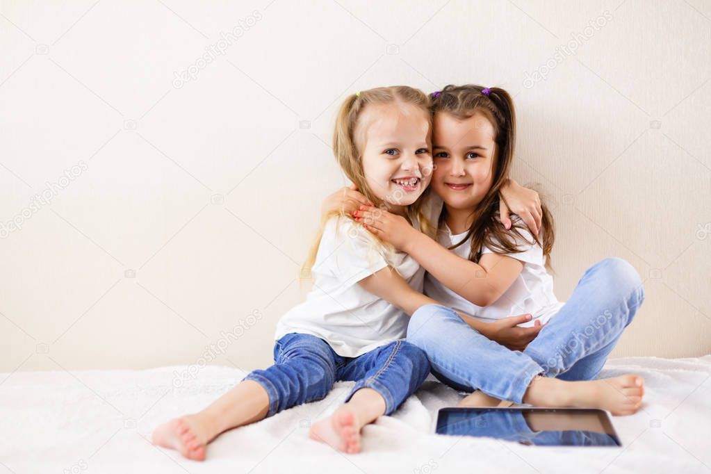 Two happy little girls hugging sitting on bed with digital tablet