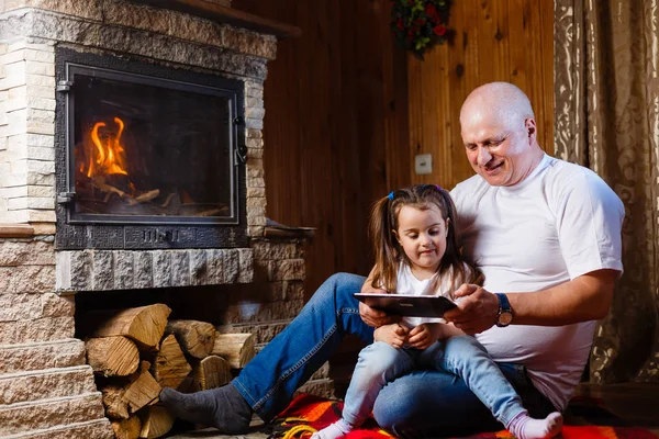 祖父と孫娘 暖炉のそばで座っているデジタル タブレットを使用して楽しい — ストック写真