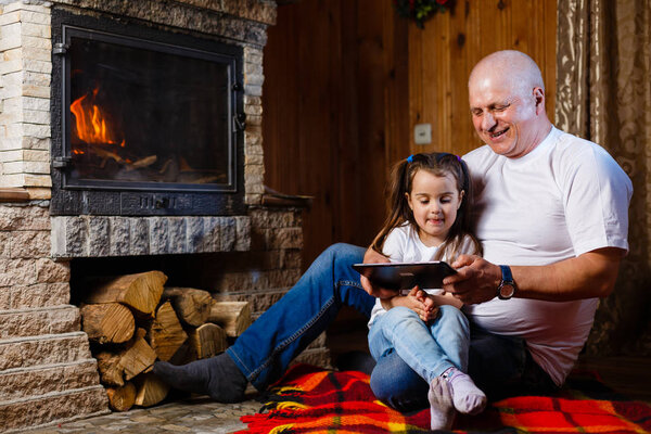 Grandfather and granddaughter having fun using digital tablet sitting at fireplace