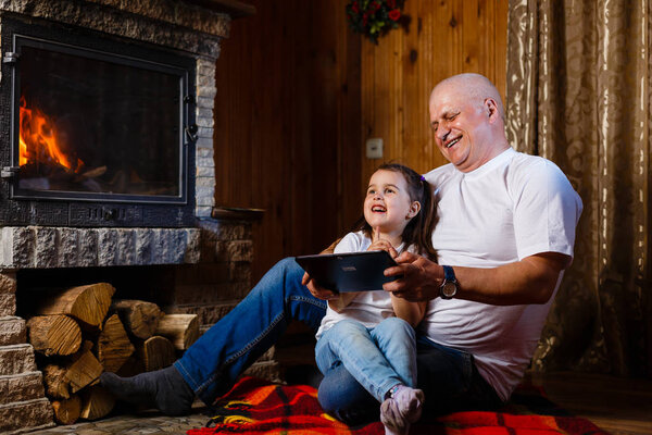 Grandfather and granddaughter having fun using digital tablet sitting at fireplace